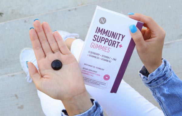 A woman in white pants and a jean jacket holding a box of the Immunity Support+ Gummies in her right hand and Immunity Support+ Gummy in the left hand.
